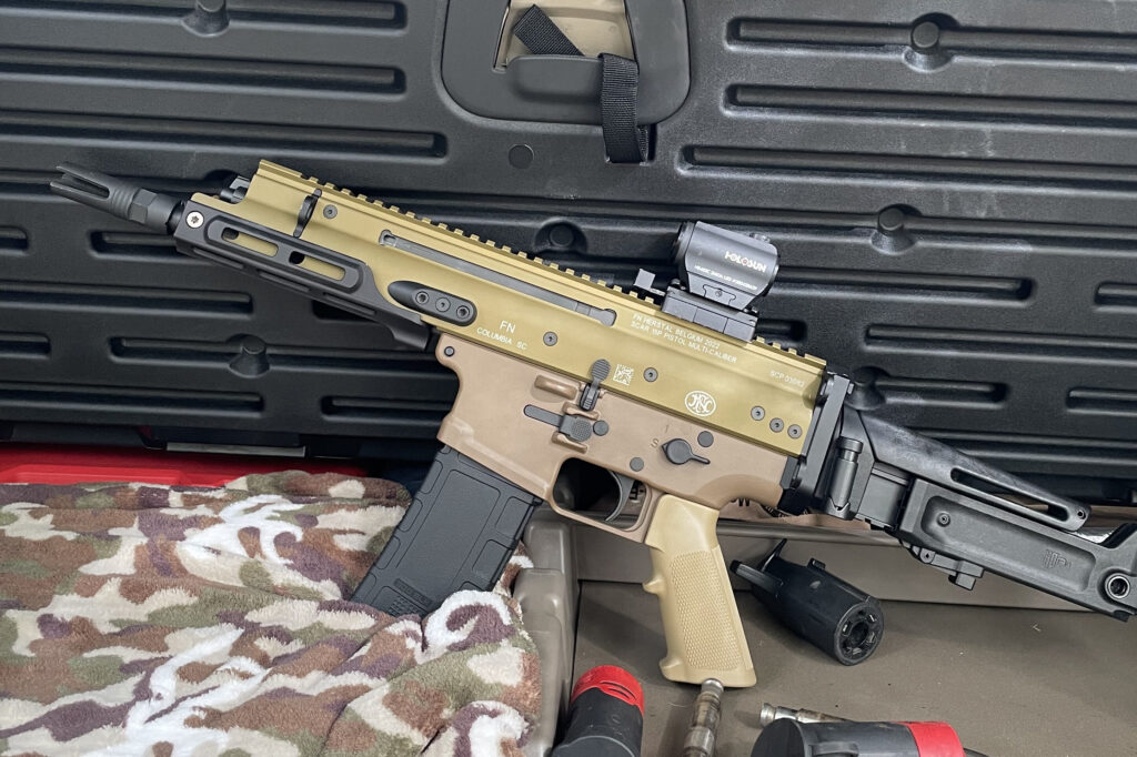 A close-up of a SCAR 15P pistol with a red dot optic, positioned on a camo cloth. The firearm is in front of a textured black gun case, with tools and accessories visible in the foreground.