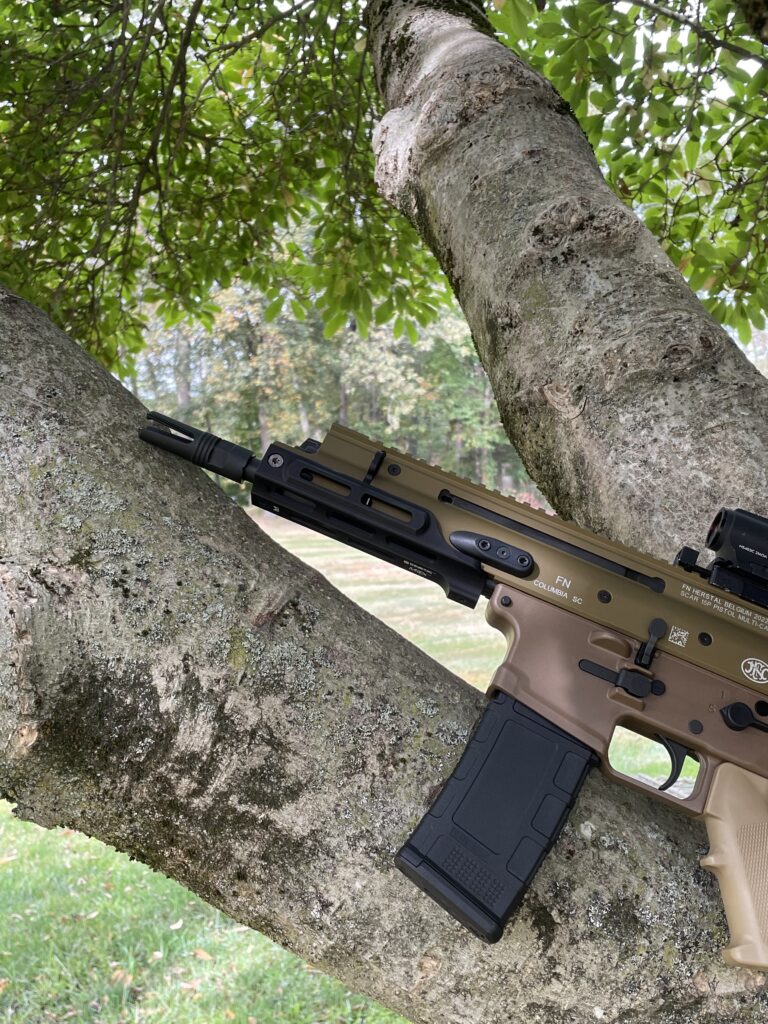 Close-up of an FN SCAR 15P firearm propped against a tree with textured bark, under a canopy of green leaves. The rifle features a two-tone design and visible optics, blending into the outdoor environment.