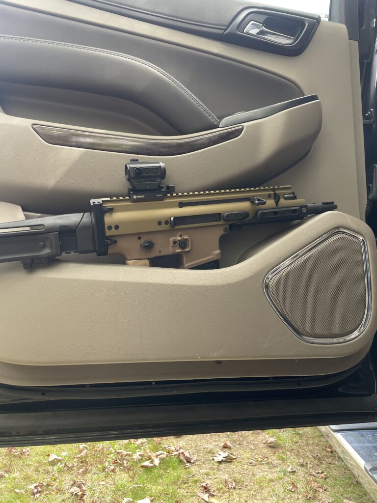 Side profile of an FDE FN SCAR 15P pistol nestled into the tan interior door panel of a truck. The door is open, with a view of the ground and leaves outside.