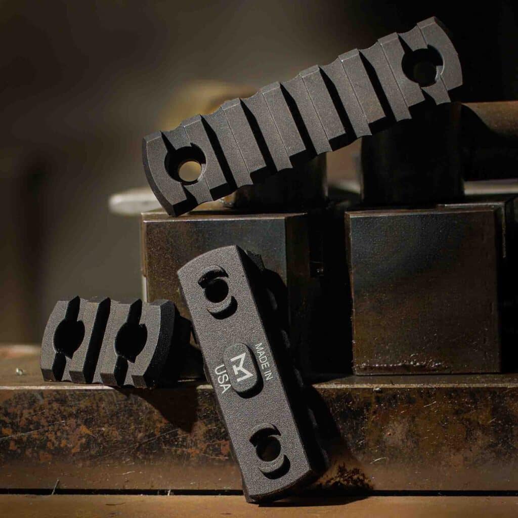 Black aluminum M-LOK Picatinny rail sections displayed on industrial metal blocks, featuring a 7-slot rail, a 3-slot rail, and an underside view showing the M-LOK logo and 'Made in USA' markings.